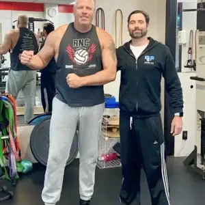 Two men posing together inside a personal training gym, one flexing his arm.