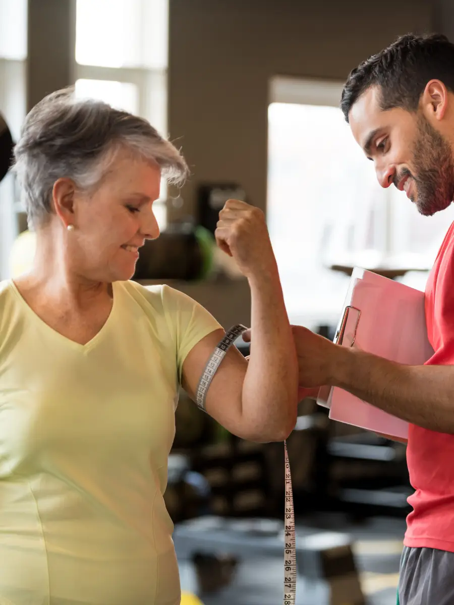 Personal trainer taking arm measurement on older female client inside a gym.