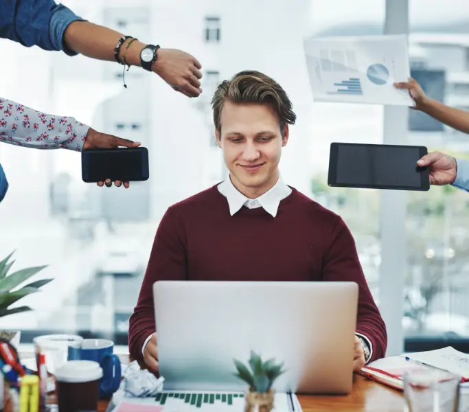 Man working on laptop while surrounded by multiple hands holding devices and documents.