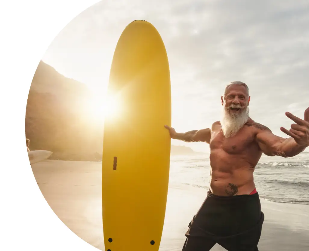 Active older man holding a surfboard on the beach at sunset, representing healthy aging and vitality.