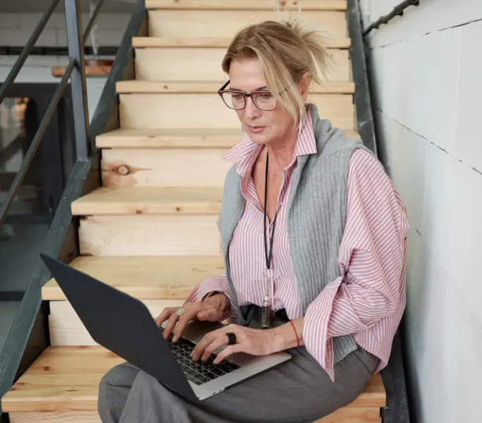 Woman sitting on stairs working on a laptop with focused expression.